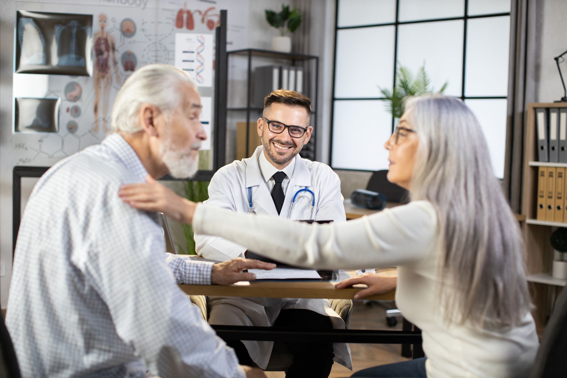 Doctor examining an older patient while a woman comforts him in a clinic office