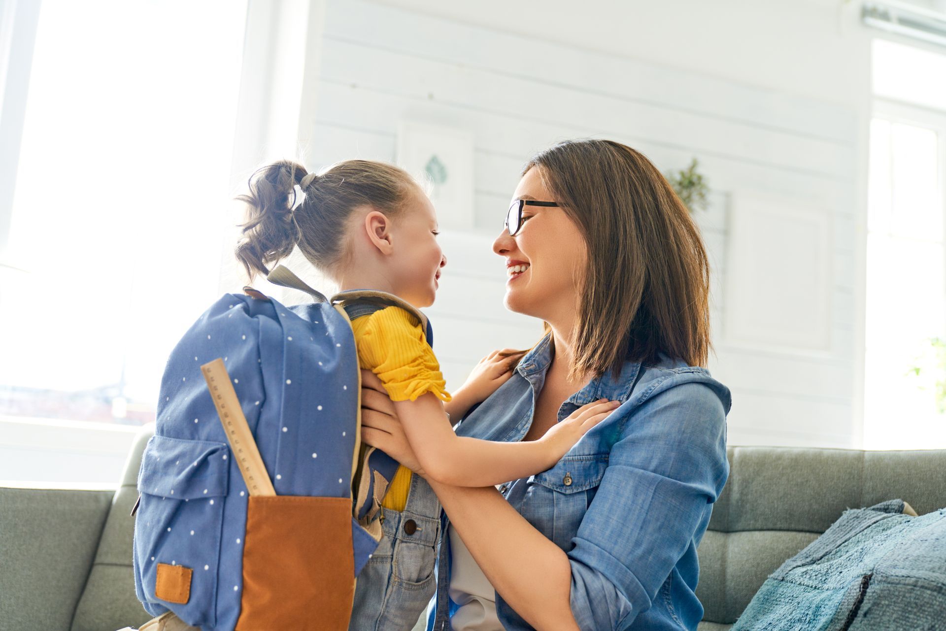 Two people smiling and embracing on a couch in a bright living room, with a backpack nearby