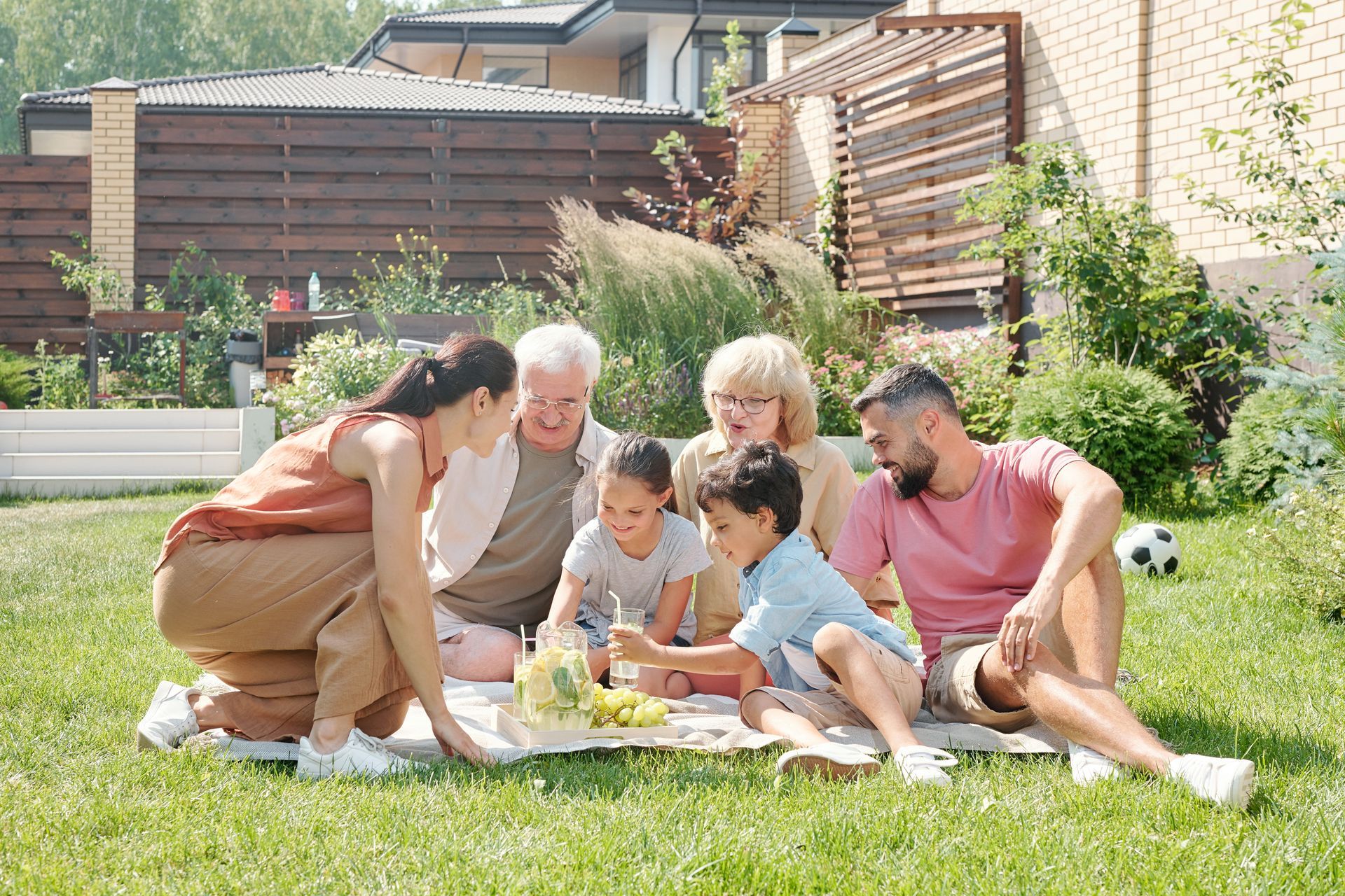 Family sitting on a blanket in a sunny backyard, sharing a picnic and looking at a tablet.
