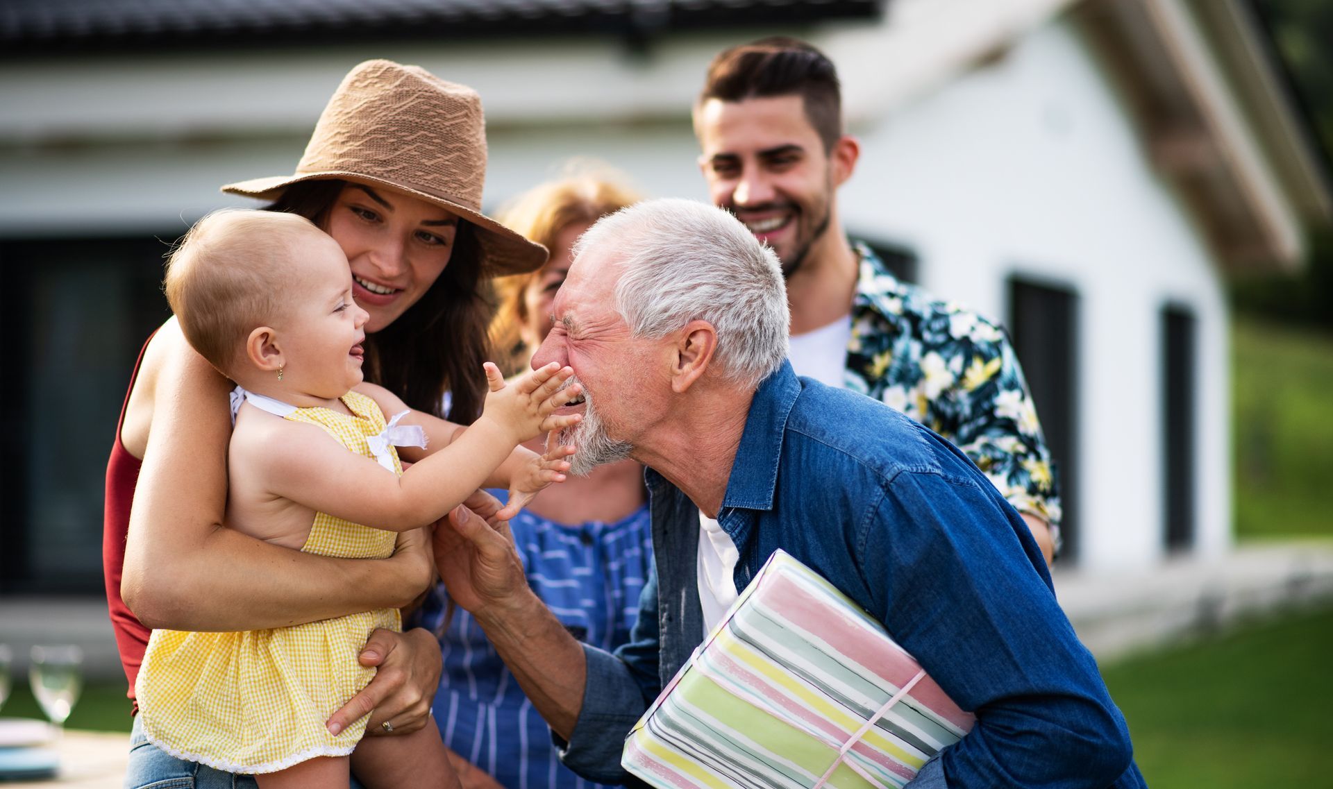Family outdoors in a yard, an older man smiling at a baby while a woman holds the child and a man stands behind.