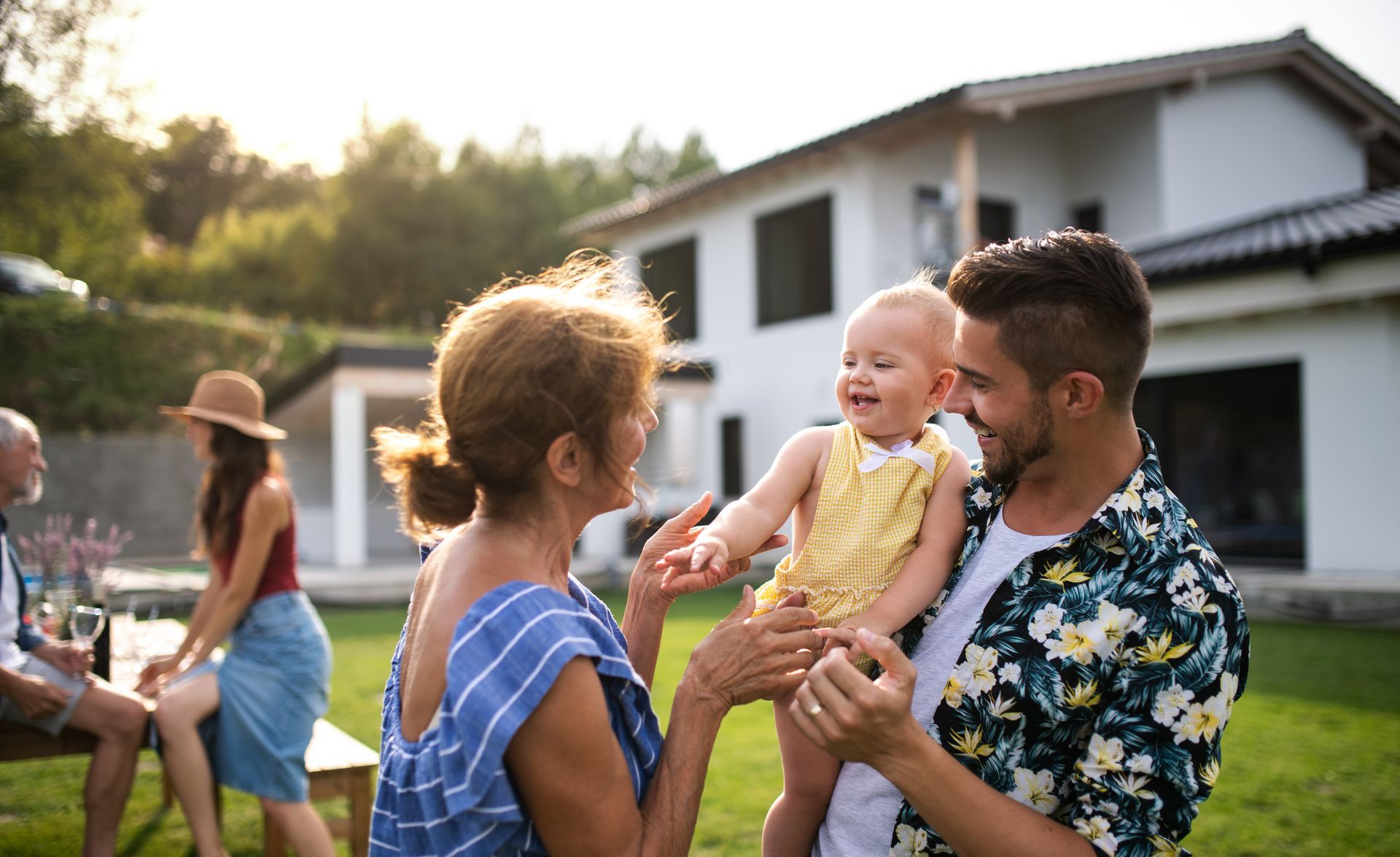 Man holding a smiling baby in a yard, with a child and a woman near a white house at sunset