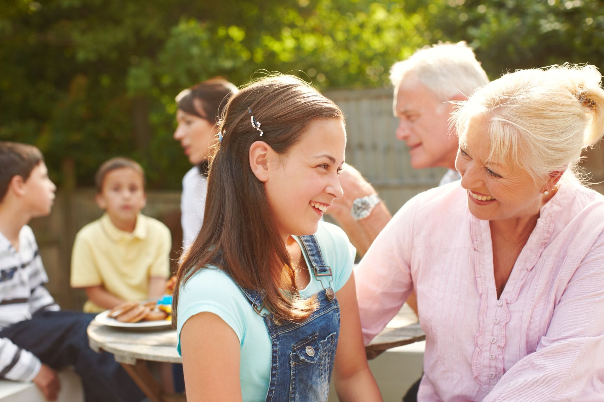 Two women smiling and talking outdoors at a sunny community gathering with children in the background