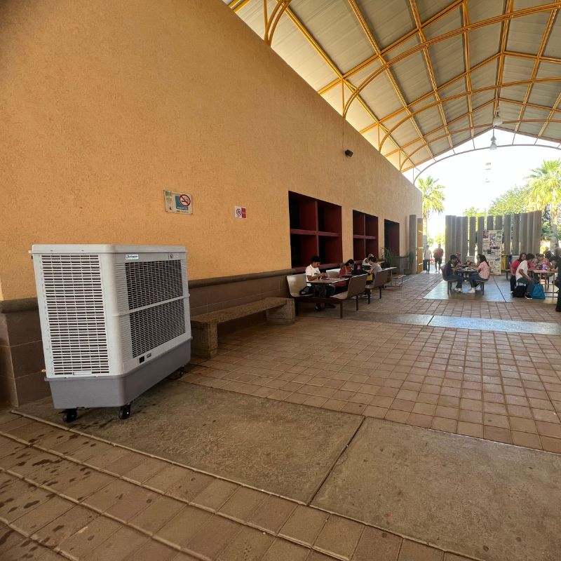 Un grupo de personas están sentadas en mesas en un pasillo grande, del lado izquierdo un aerocooler portátil.