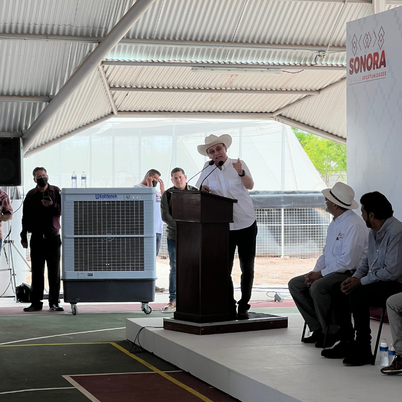 El gobernador de Sonora Alfonso Durazo está de pie en un podio dando un discurso de fondo un aerocooler portátil.