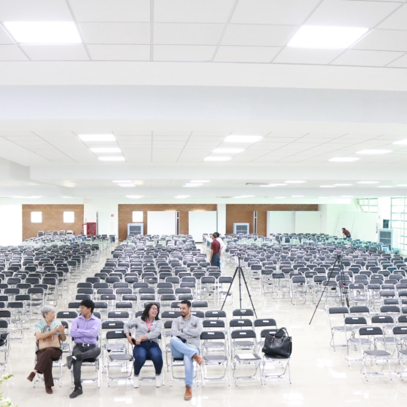 Un grupo de personas están sentadas en filas de sillas, de fondo varios enfriadores evaporativos portátiles.