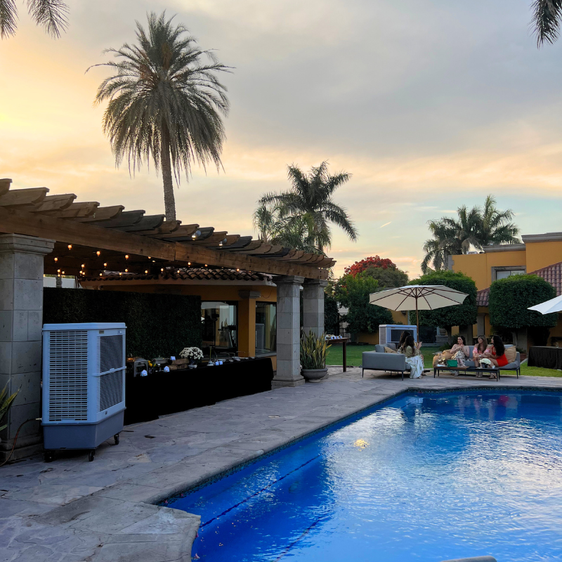 Una gran piscina rodeada de palmeras y sombrillas y en un costado un aerocooler portatil.