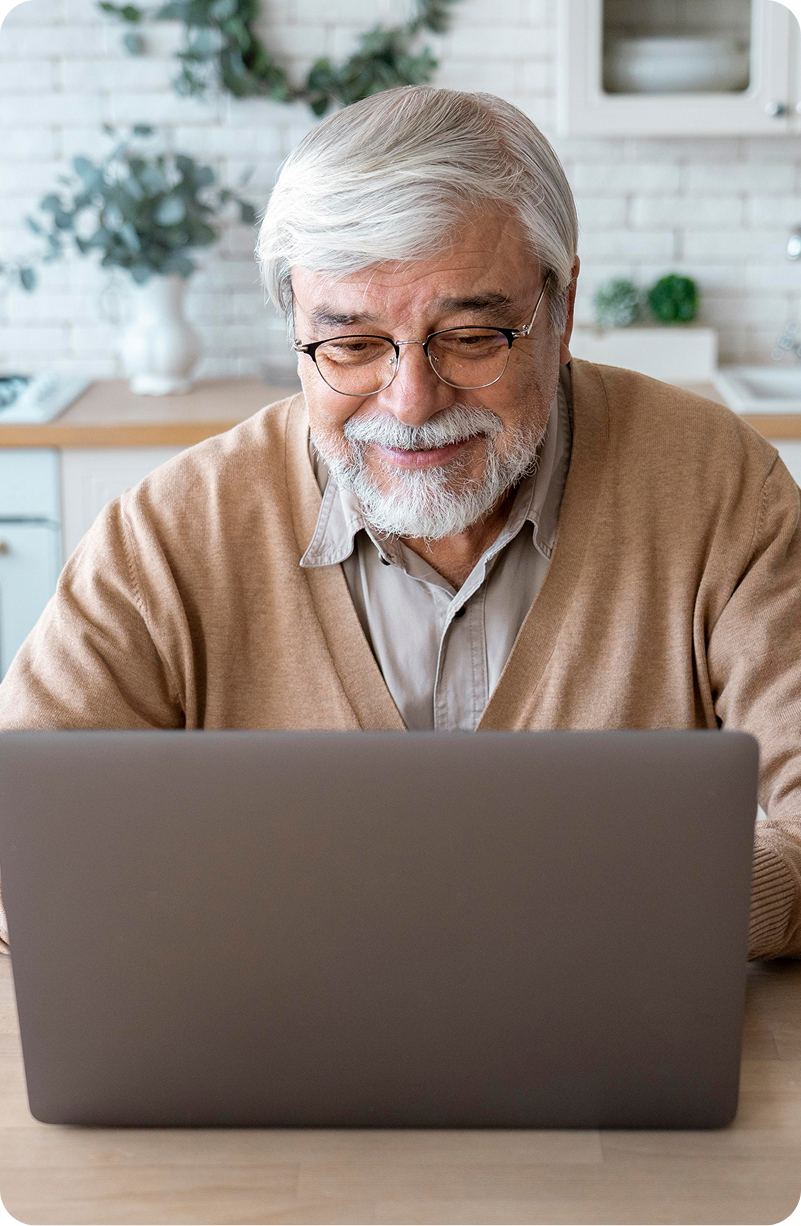An elderly man is sitting at a table using a laptop computer.