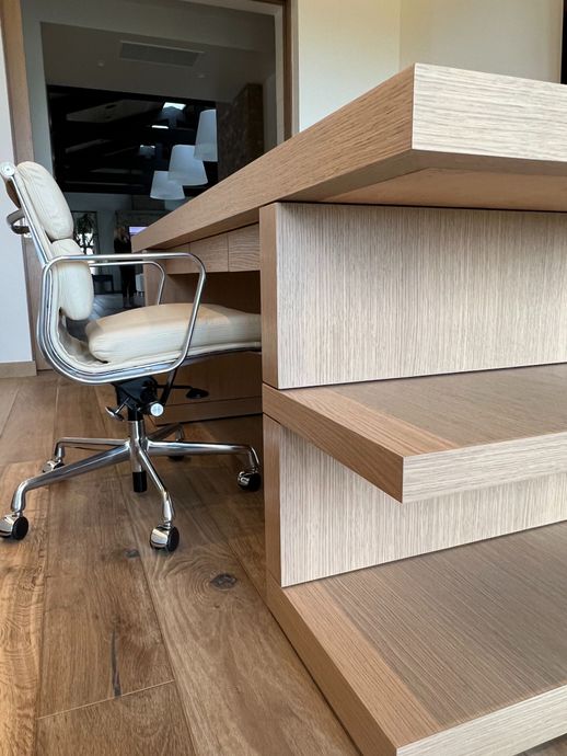 A light-wood desk with a tiered, stepped-base design sits on a wooden floor next to a white office chair.