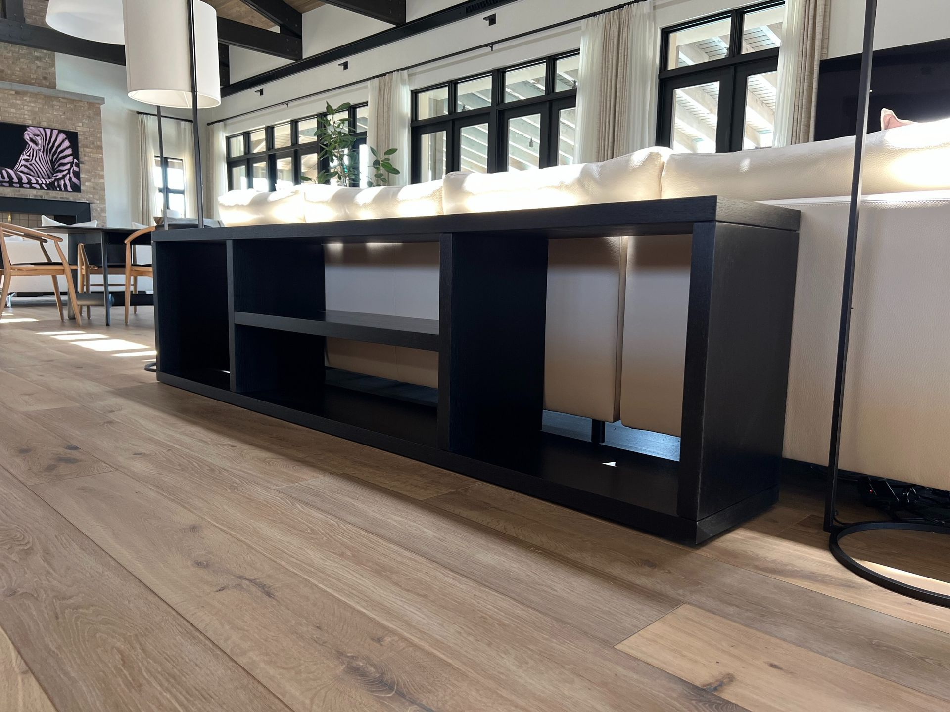 A black, low-profile storage console sits on light wood flooring behind a white sofa in a bright, modern living room.