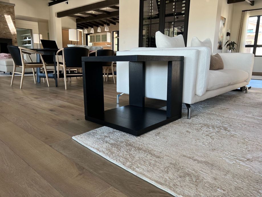 A modern black side table sits on a light-colored rug next to a white sofa in a contemporary, sunlit living room.