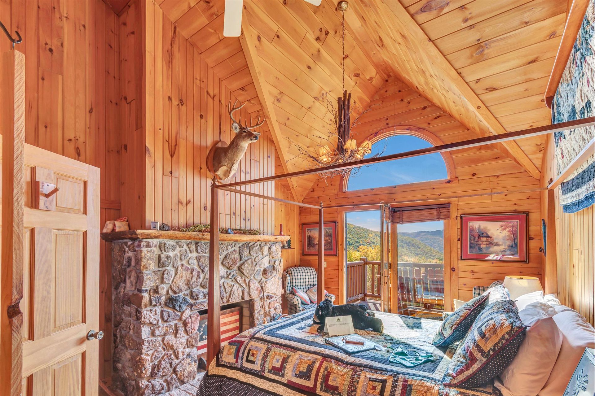 Cozy wooden bedroom with four-poster bed, stone fireplace, and mountain view through a curved window and open doors.