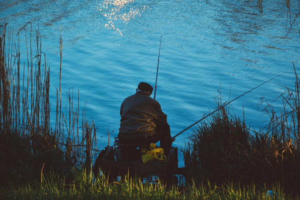 Person fishing by a body of water at dusk, holding a fishing rod.