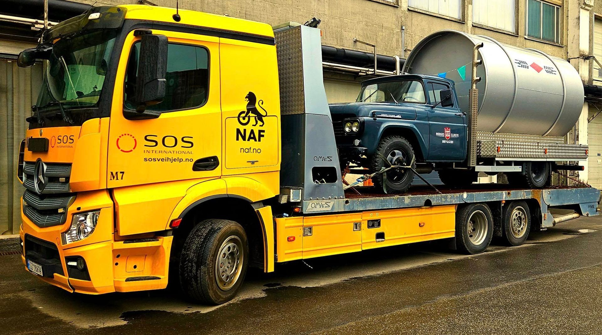 Gul bergingsbil som frakter et blått kjøretøy med en stor sølvfarget tank på lasteplanet, parkert i en bygate.