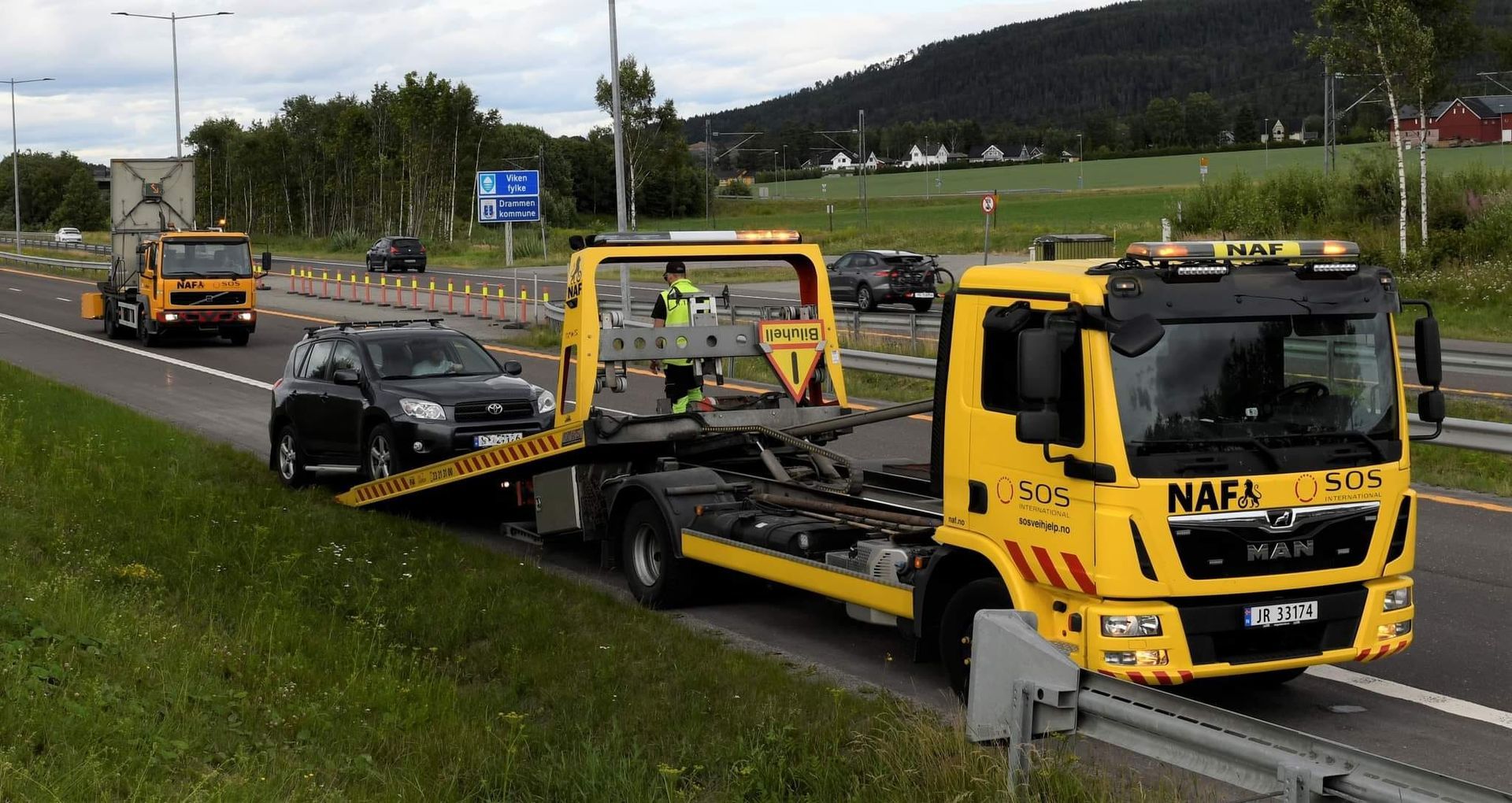 Gul bergingsbil laster en svart bil på siden av en motorvei, med en annen bergingsbil i bakgrunnen.