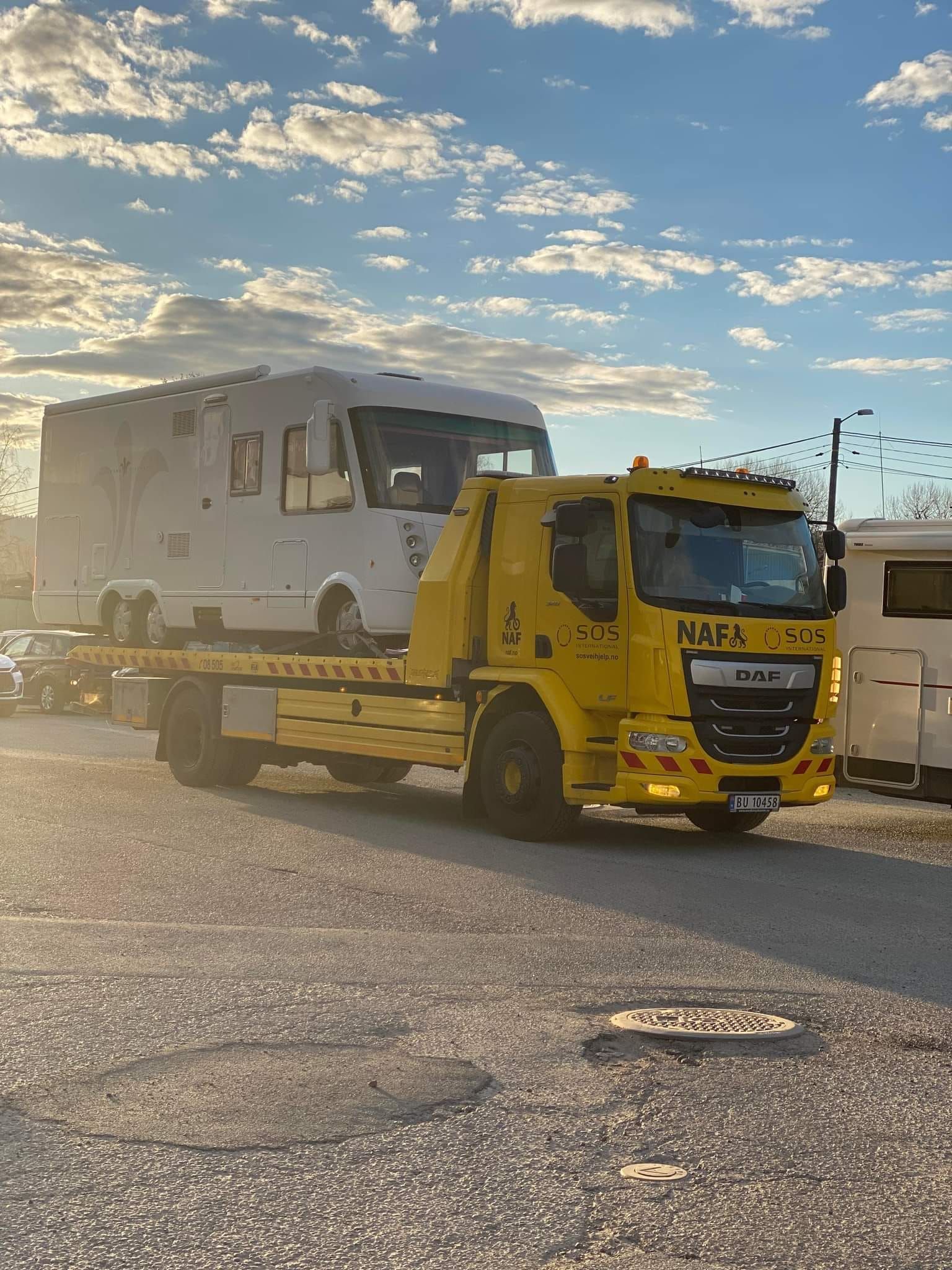 En gul bergingsbil som frakter en hvit bobil på en parkeringsplass, med en blå himmel i bakgrunnen.