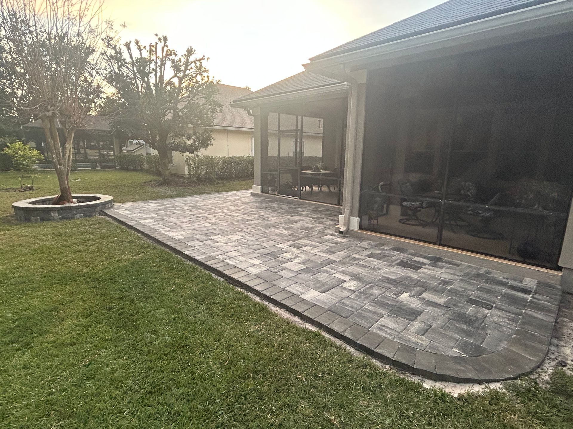 A rectangular gray paver patio extends from a screened-in porch into a grassy backyard with a small circular tree planter.