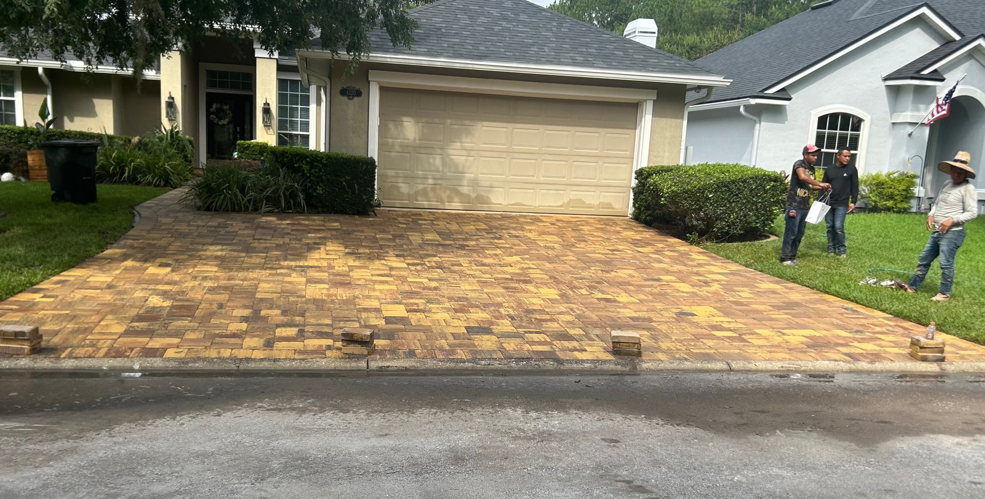 Three workers install new tan and brown stone pavers on a driveway in front of a residential garage.