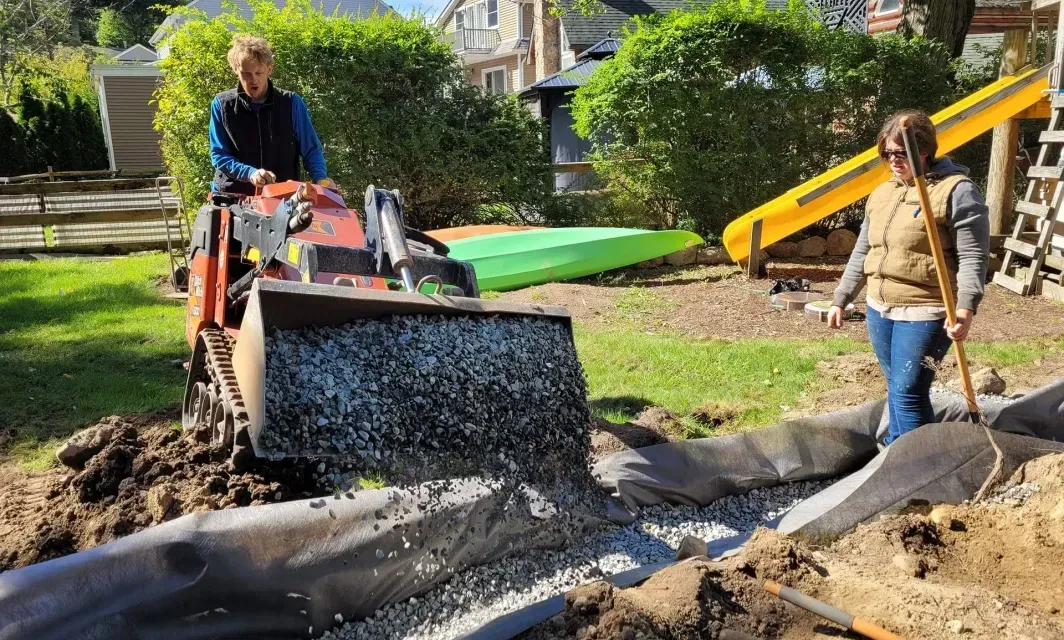 A man is driving a bulldozer in a yard while a woman stands next to him.