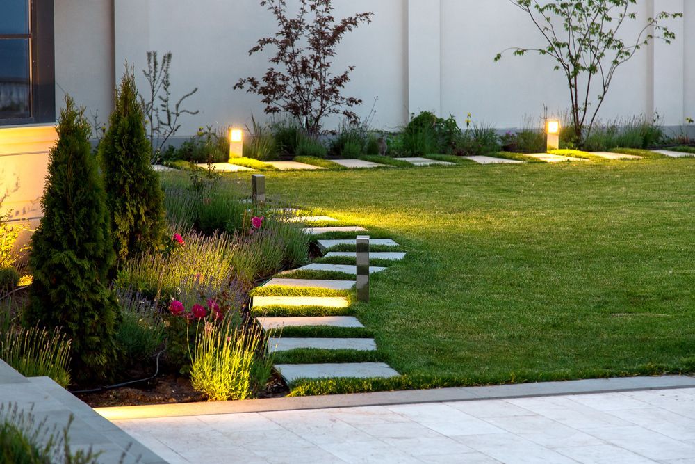 Stone path lit by lights leads through a green lawn and garden with small trees.
