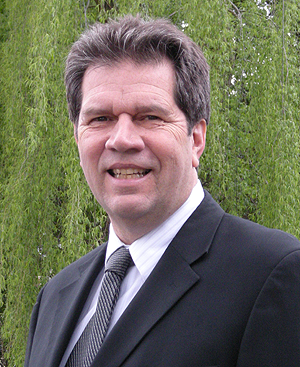 Man in a dark suit smiles, outdoors near green foliage.
