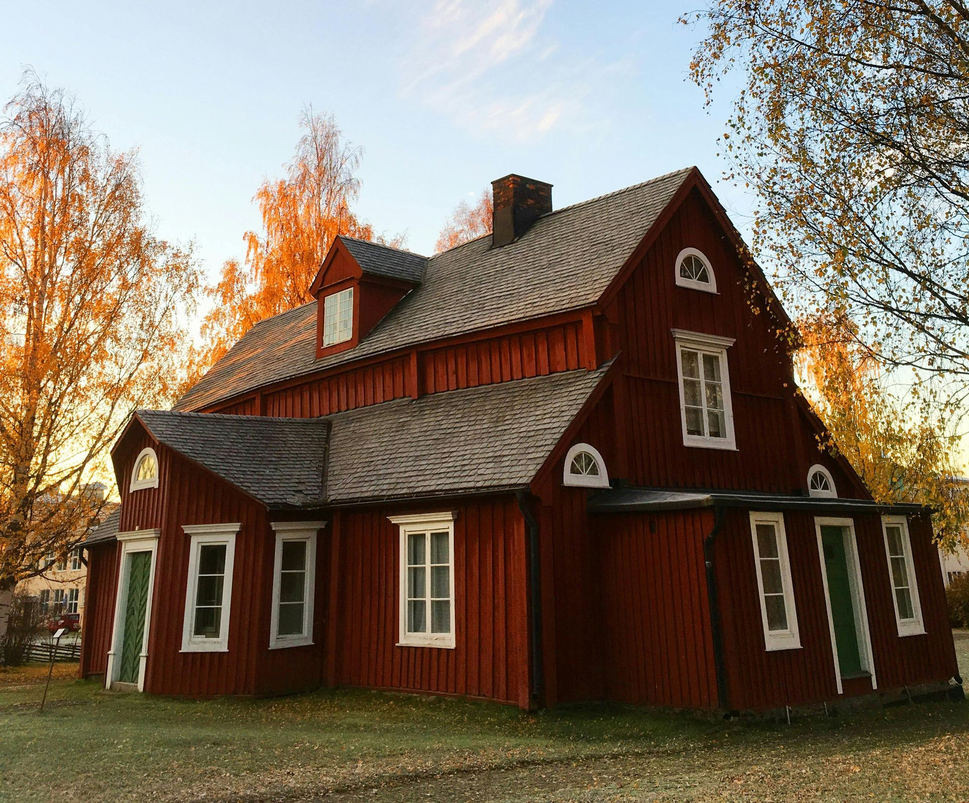 Red wooden building with white-trimmed windows and a dark roof, surrounded by trees with autumn foliage.
