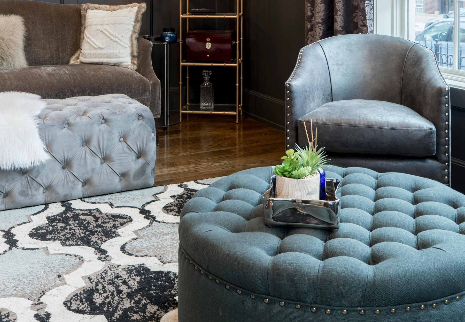Living room with grey tufted ottoman and armchair, patterned rug, sofa and gold shelving.