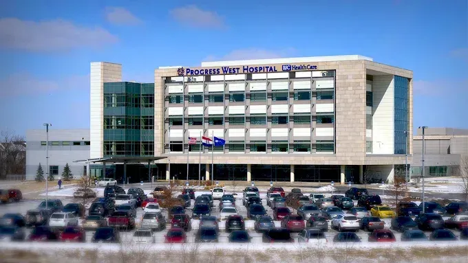 Hospital building exterior with many parked cars and flags on a sunny day.