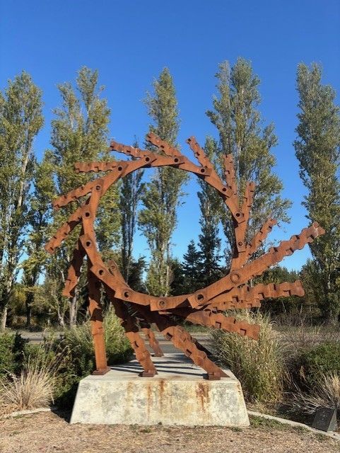 Rust-colored abstract metal sculpture on a concrete base in a grassy park with tall trees and blue sky