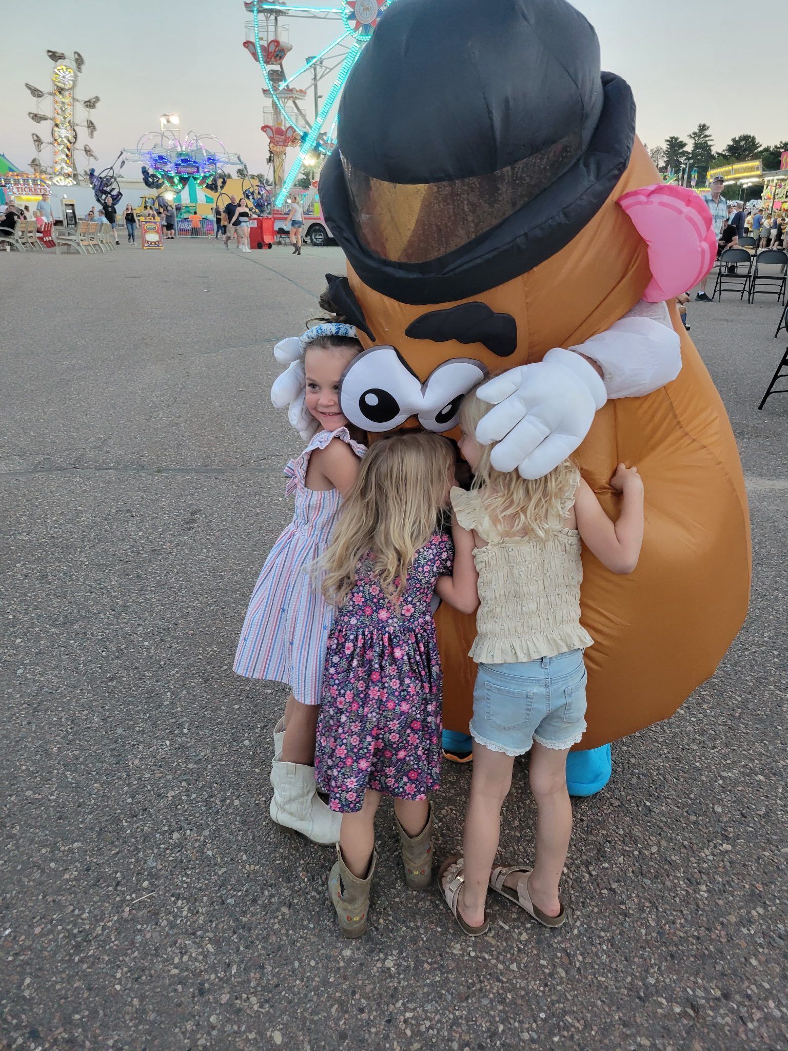 Three girls hugging a large inflatable Mr. Potato Head at a carnival with a Ferris wheel.