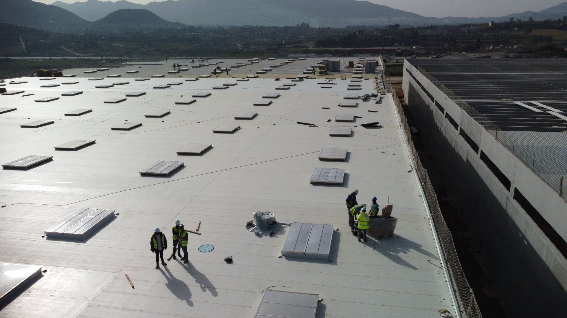 Obreros instalando materiales en un tejado grande y plano. Edificio gris y blanco con montañas al fondo.