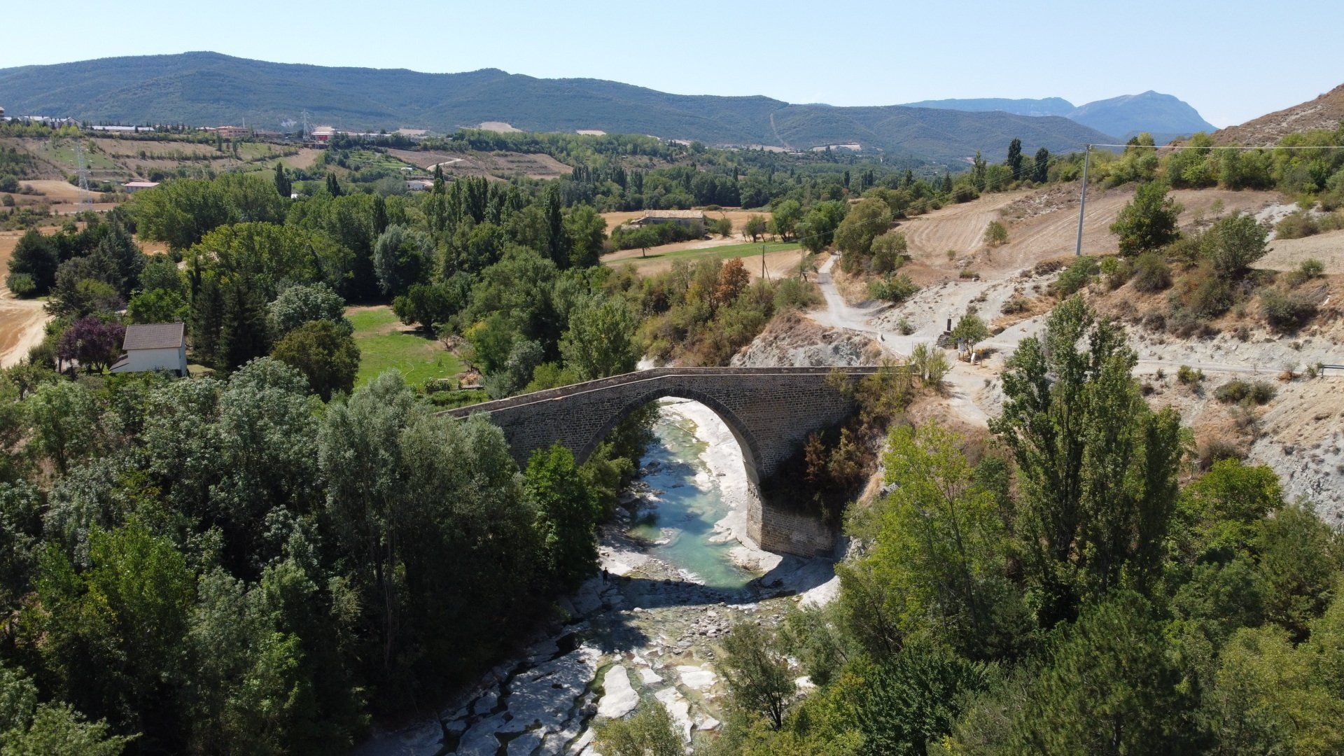 Puente de piedra sobre un río en un valle con árboles y colinas distantes bajo un cielo azul.