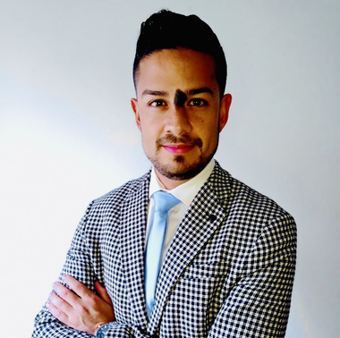 Hombre con chaqueta a cuadros, corbata azul claro, brazos cruzados, sonriendo, sobre un fondo blanco.
