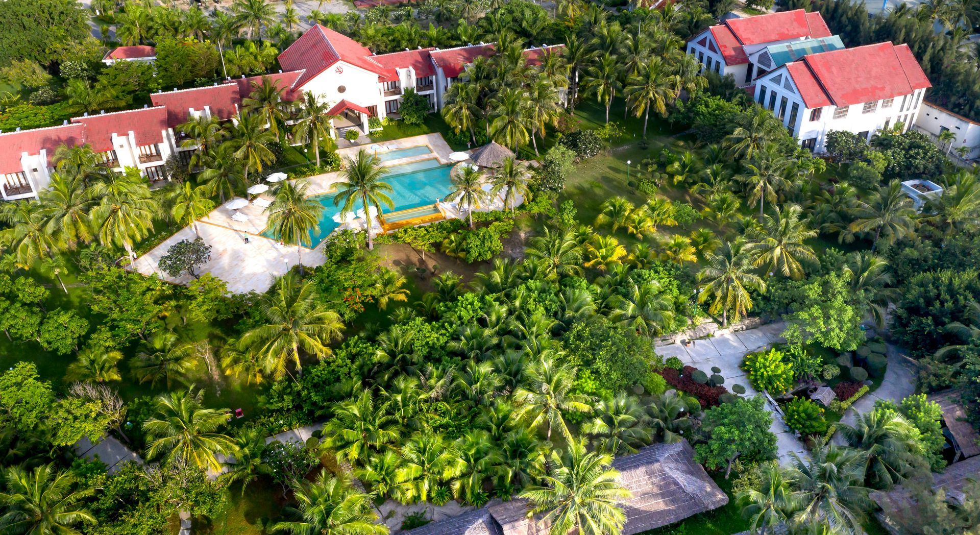 Vista aérea de un complejo turístico tropical con edificios de techos rojos, una piscina turquesa y exuberantes árboles verdes.