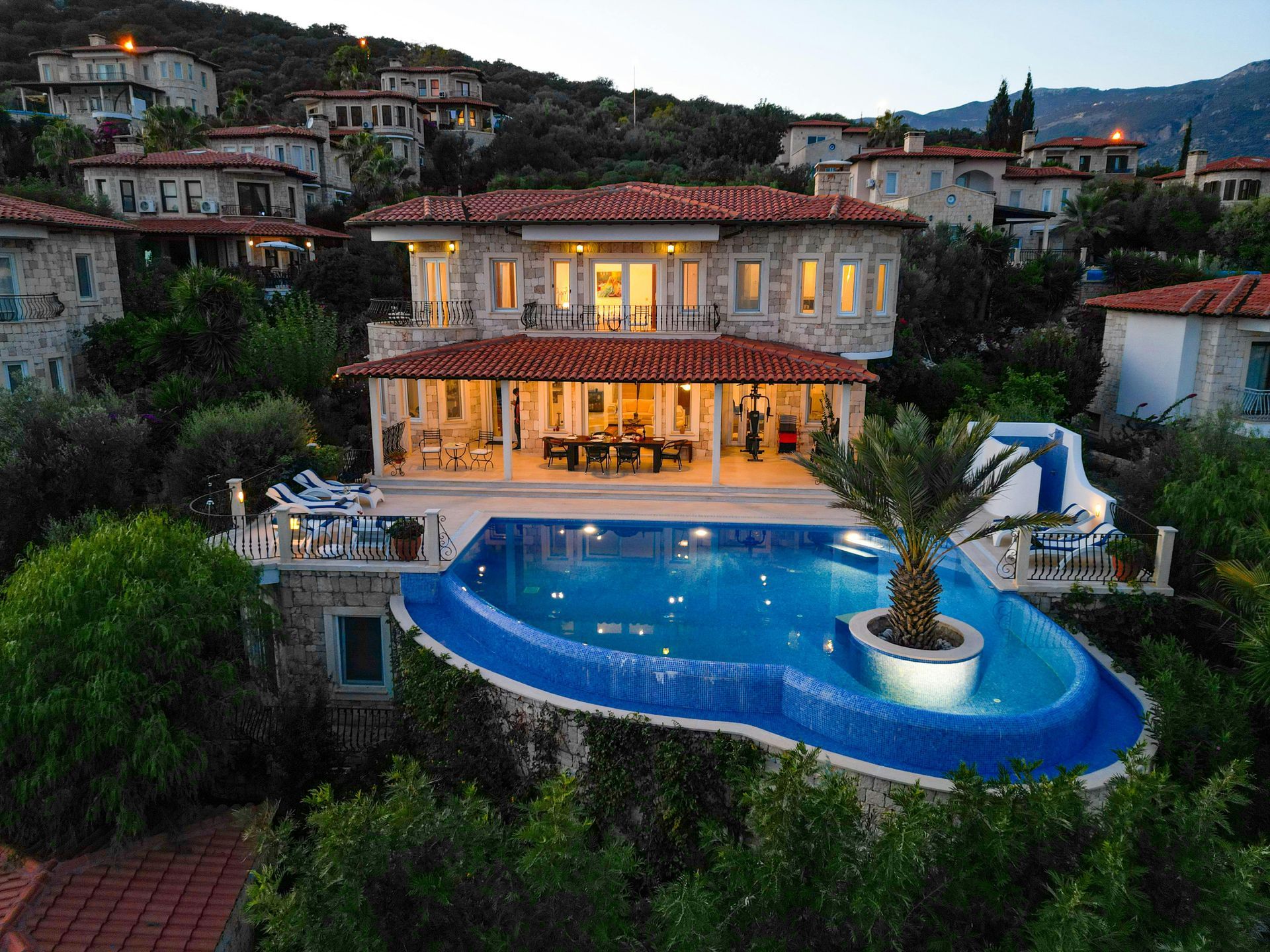 Villa de lujo en la ladera de una colina con ventanas iluminadas y una piscina azul brillante al atardecer.