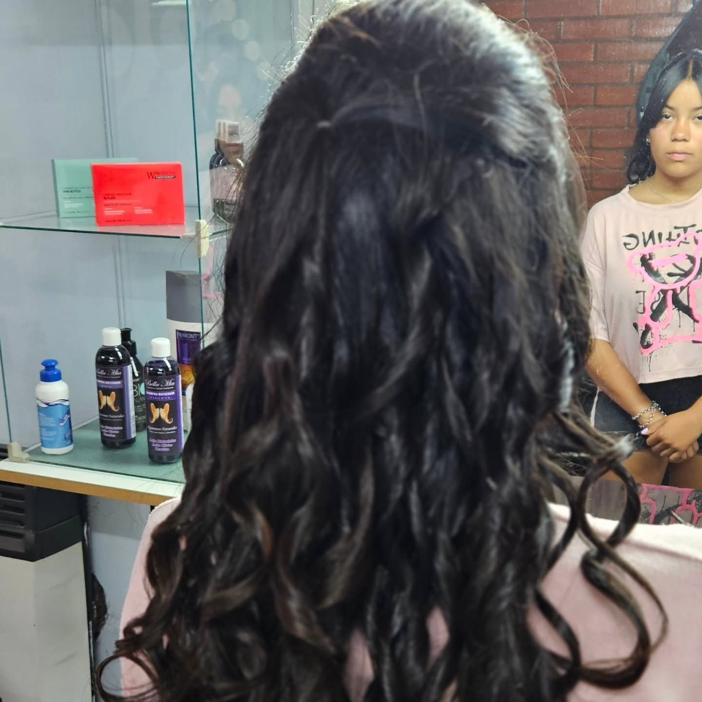 Mujer con cabello oscuro y rizado peinada en una peluquería. Otra persona se refleja en el espejo detrás de ella.