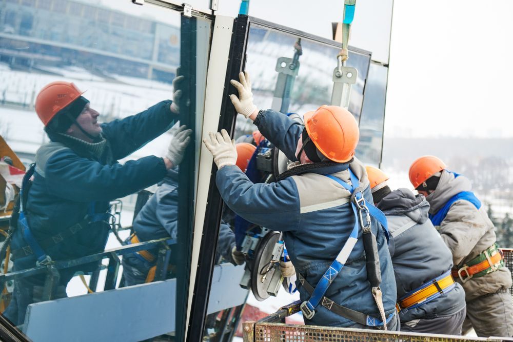 Workers in orange hard hats install large glass panel on building facade.