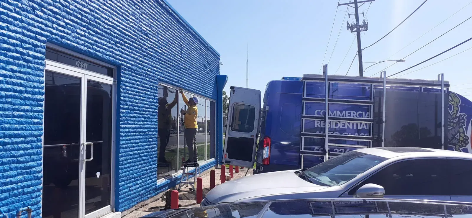 Workers cleaning windows of a blue brick building. A blue van with ladders is parked nearby.