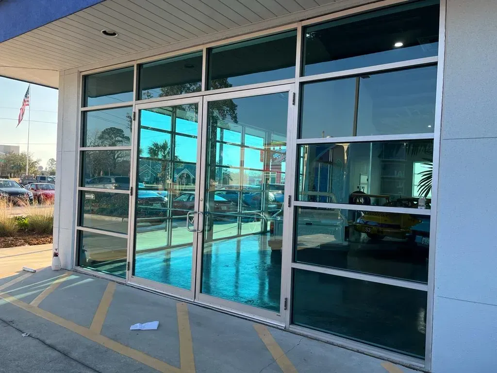 Exterior view of building with teal-tinted windows. Silver frames, reflecting blue sky, parked cars.