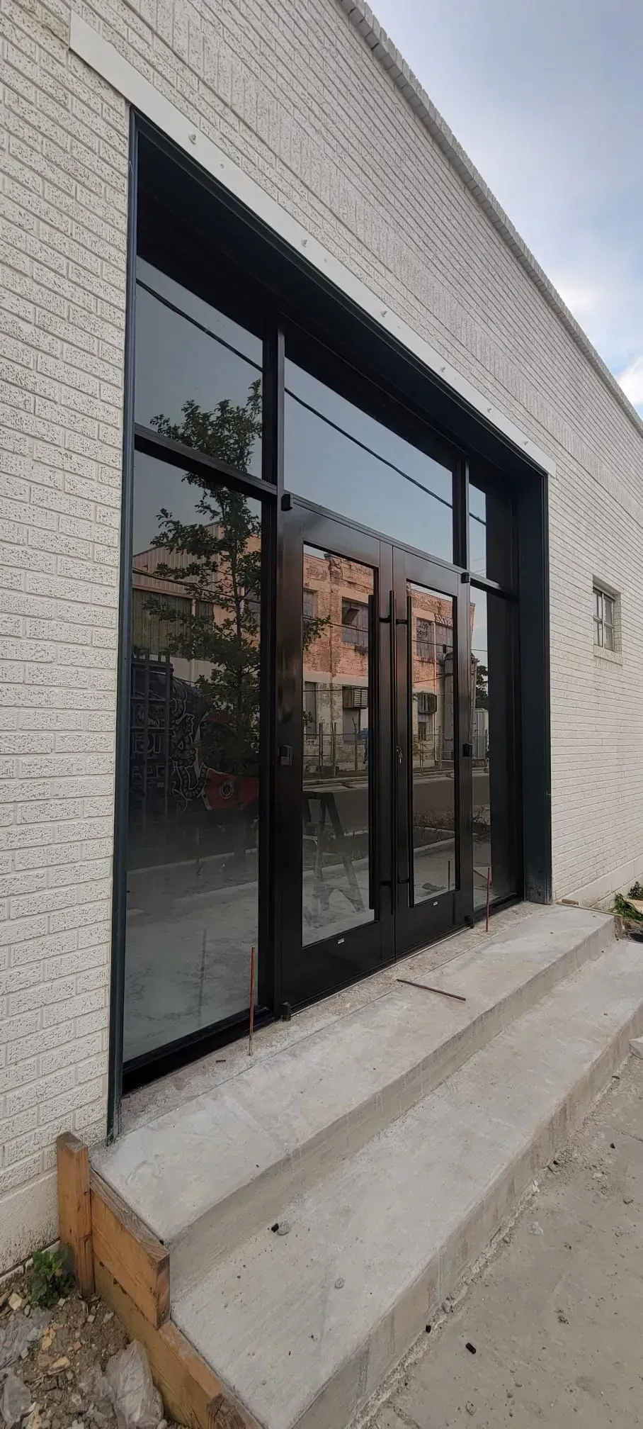 Black framed glass doors with reflection of a building and trees, over concrete steps, white brick building.
