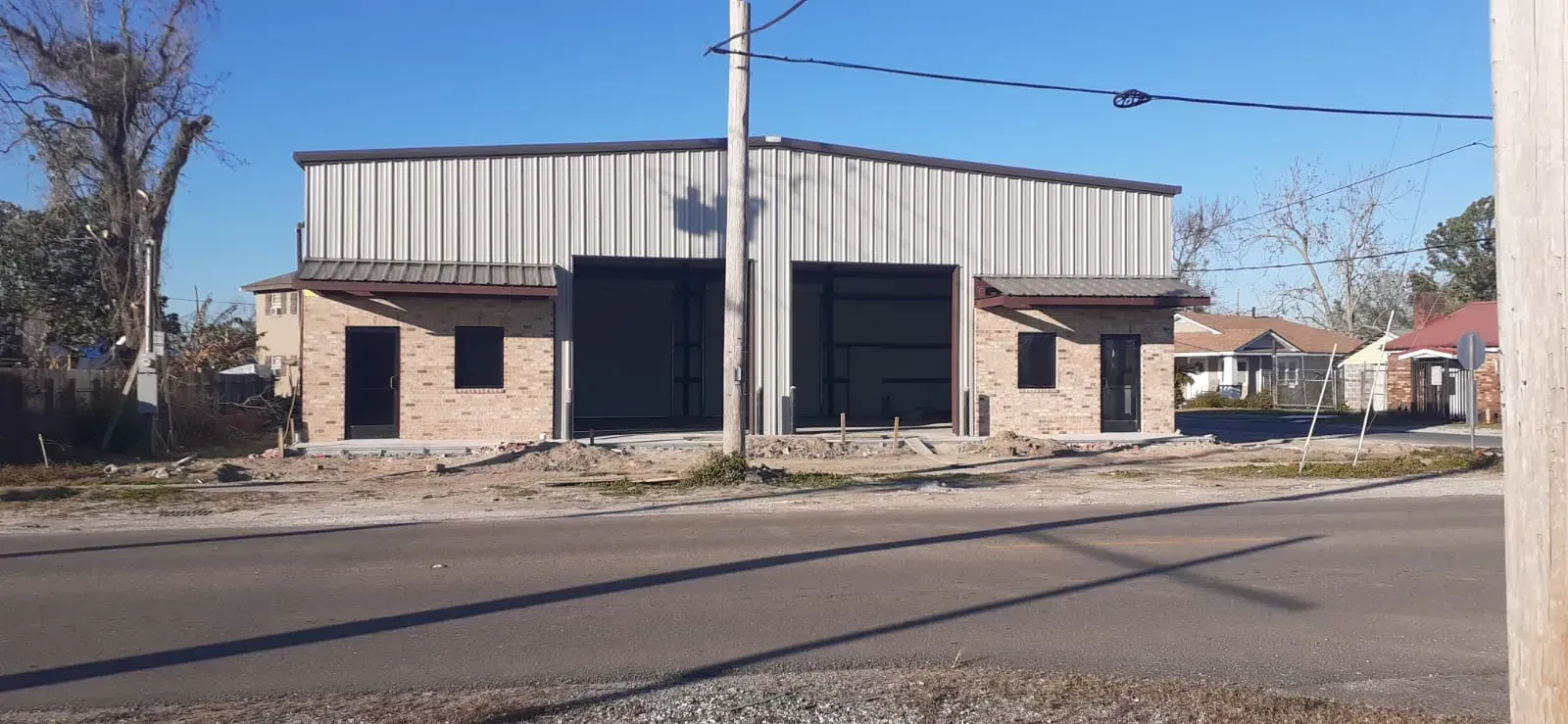 A metal-roofed building with two large garage doors and brick siding stands on a road under a blue sky.