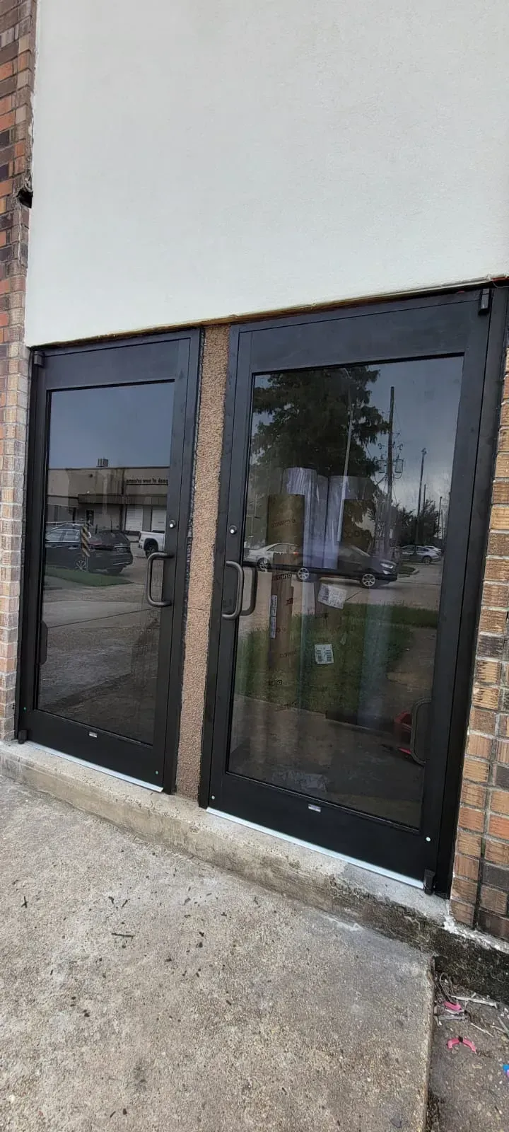 Double glass doors with black frames, reflecting street scene, set in a brick and white wall building.