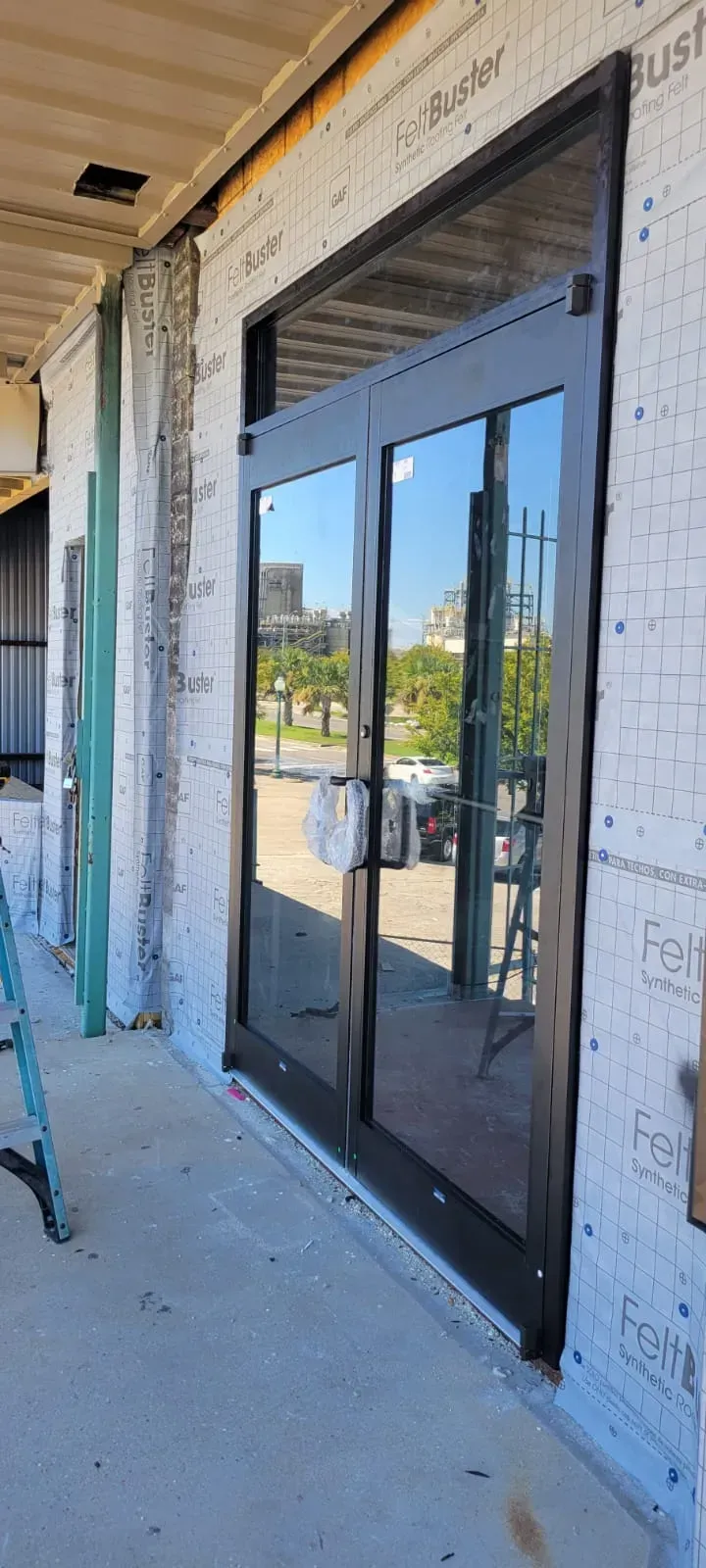 Exterior view of a building under construction, featuring black framed glass doors reflecting the outdoors.