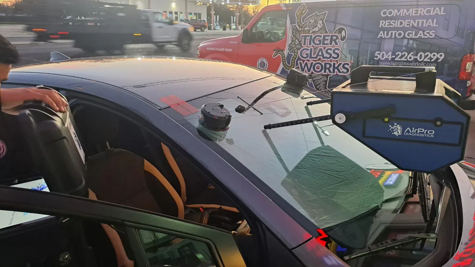 Person calibrating a car's windshield with specialized equipment; parked outdoors near traffic and a glass repair truck.