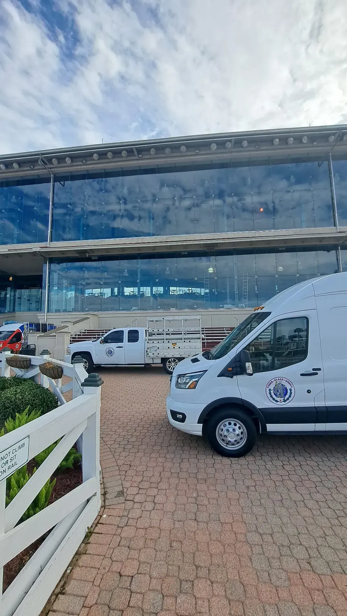 White van and truck parked near a glass-walled building with a blue sky overhead. A white fence is in the foreground.