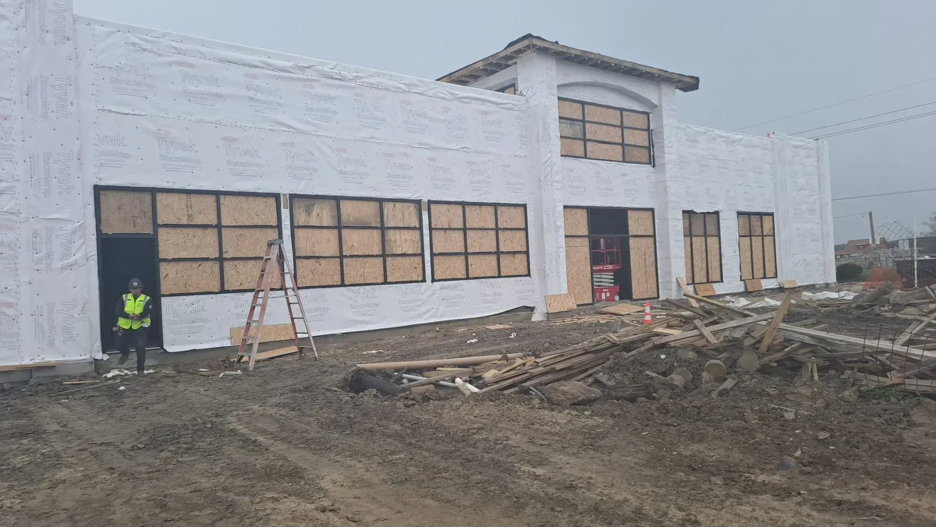 Building under construction, windows boarded. Construction worker in a safety vest stands outside the entrance.