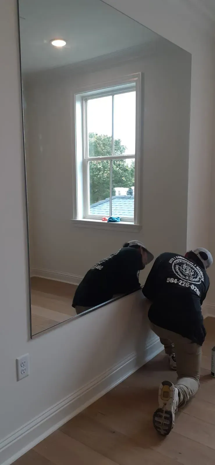 Two workers installing a large mirror on a wall in a room with a window.
