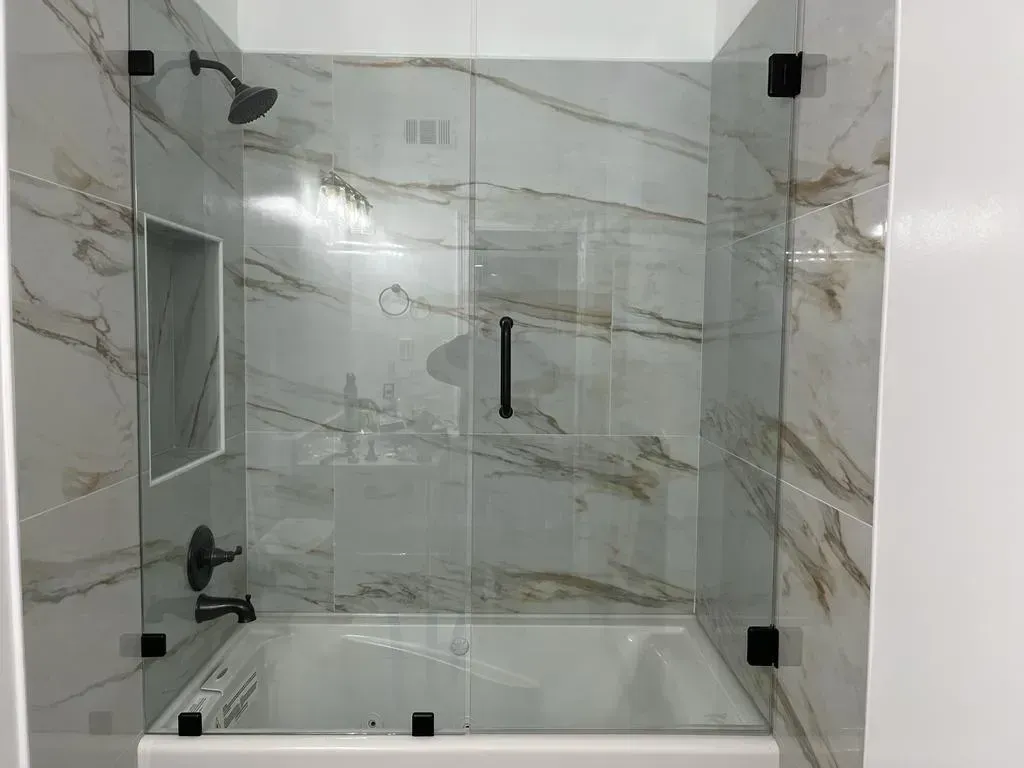 Shower with glass doors and black fixtures, marble-look tile walls, white tub.
