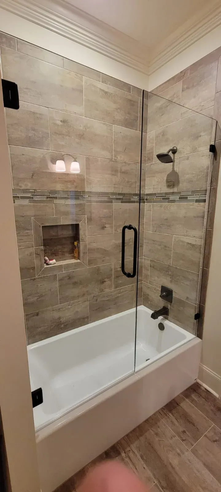 Bathroom with a white bathtub and glass shower doors, with wood-look tile on the walls and floor.