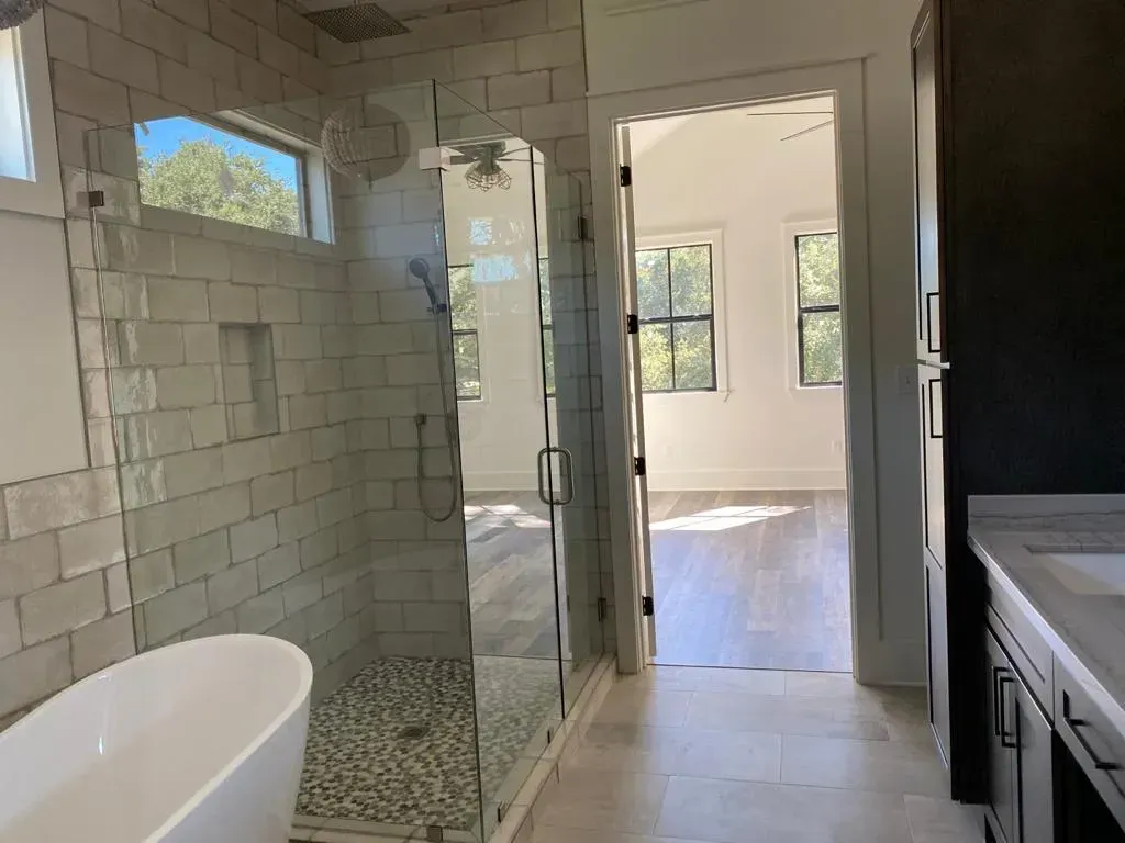 Modern bathroom with a glass shower, white tub, and doorway to a bedroom with bright windows.