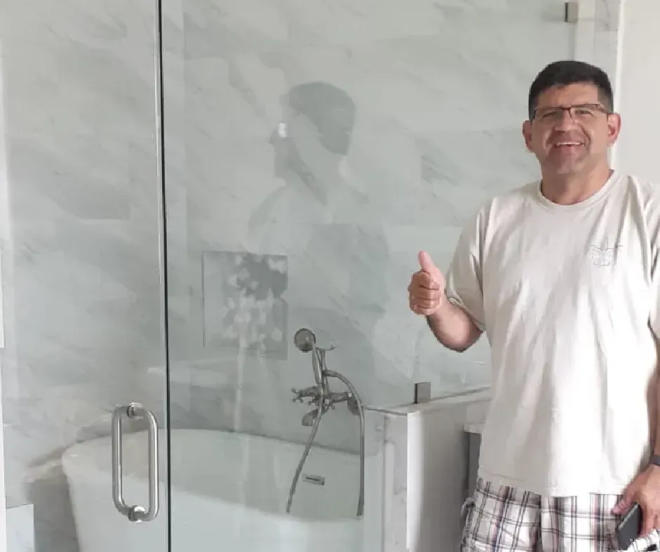 Man gives a thumbs-up in a newly renovated bathroom with a glass shower and marble-look wall tiles.